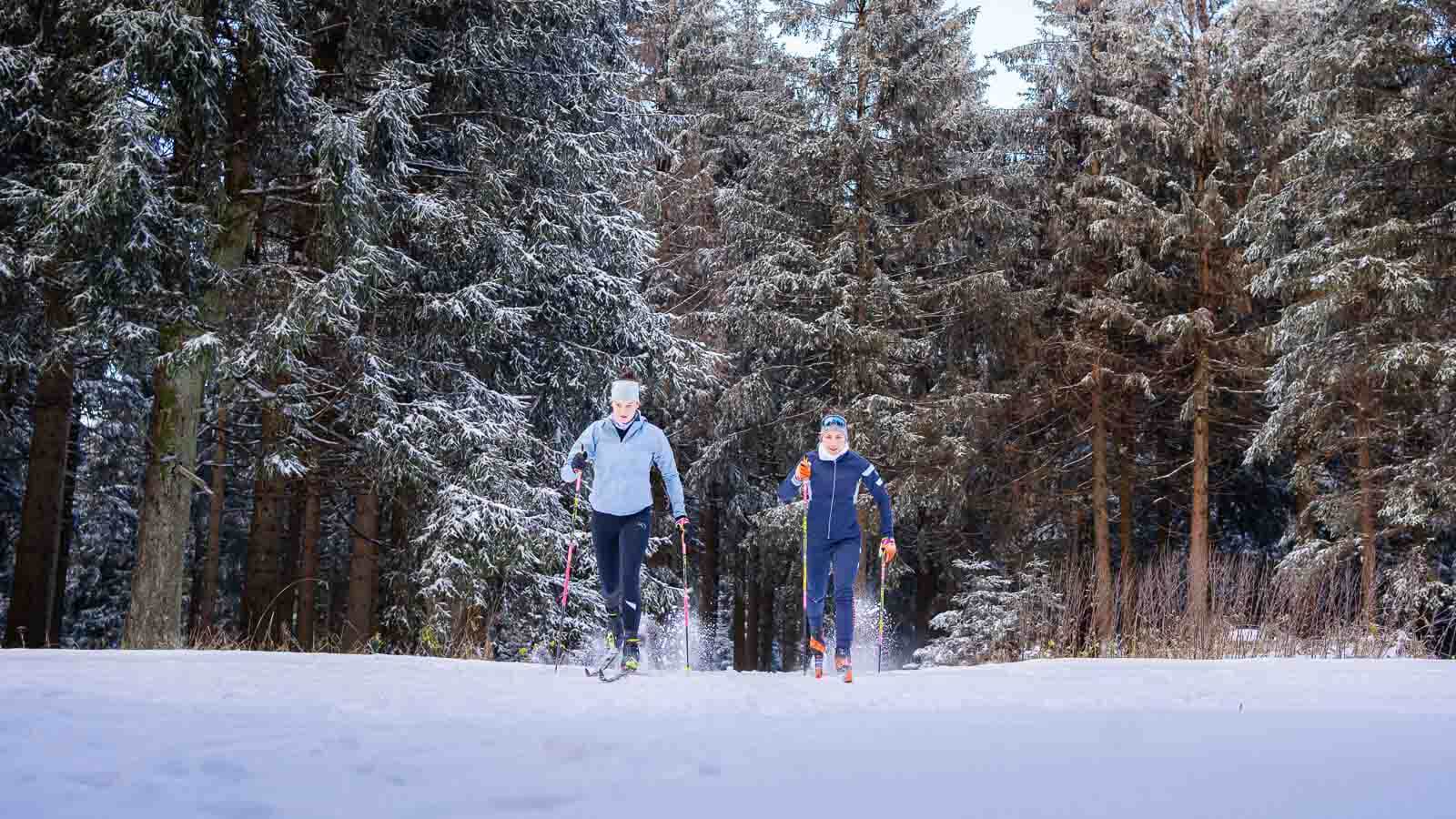 Zwei Personen laufen beim Skilanglauf durch eine verschneite Landschaft. Beide tragen warme Funktionskleidung, Wintermützen und nutzen Skistöcke. Die Umgebung ist geprägt von verschneiten Bäumen und einer klaren Winteratmosphäre.
