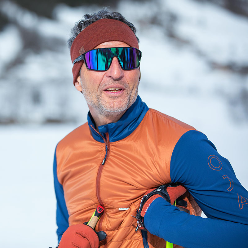 Ritratto di uno sciatore nella foresta invernale Un uomo anziano con occhiali da sci, una fascia marrone e una giacca invernale arancione-blu è stato fotografato in un ritratto davanti a un bosco bianco.