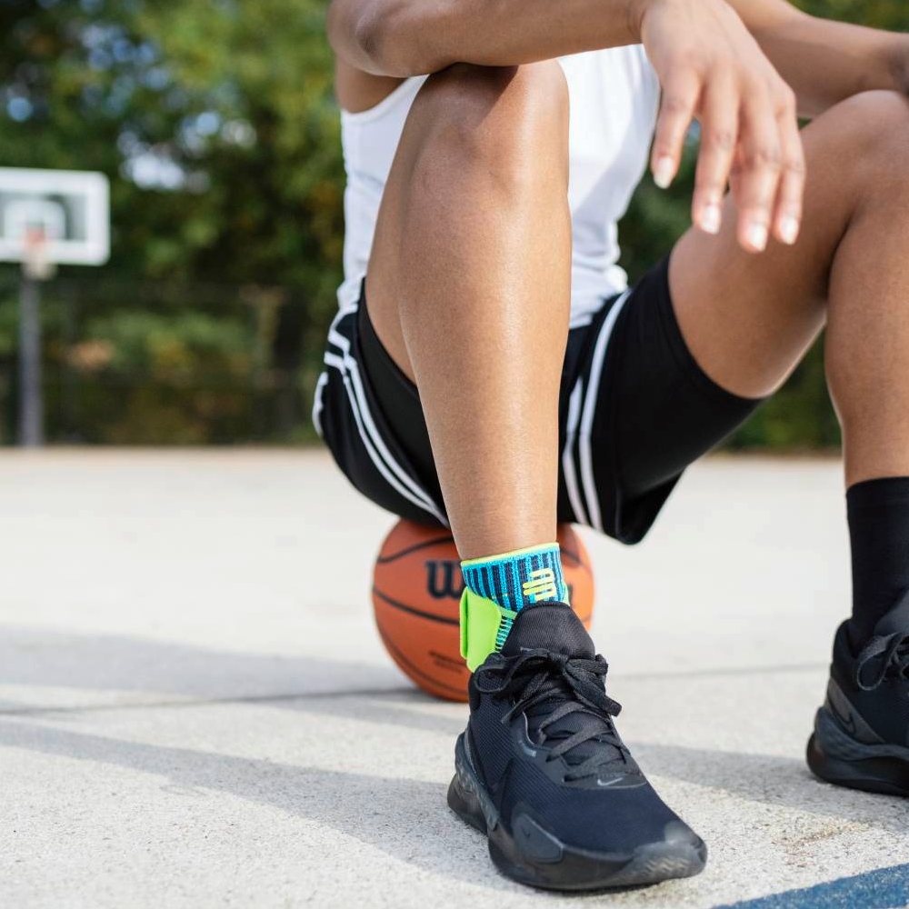 Un giocatore di basket è seduto sul campo con un pallone sotto la gamba. Indossa la Bauerfeind Sports Ankle Support Rivera in blu-verde sulla caviglia.