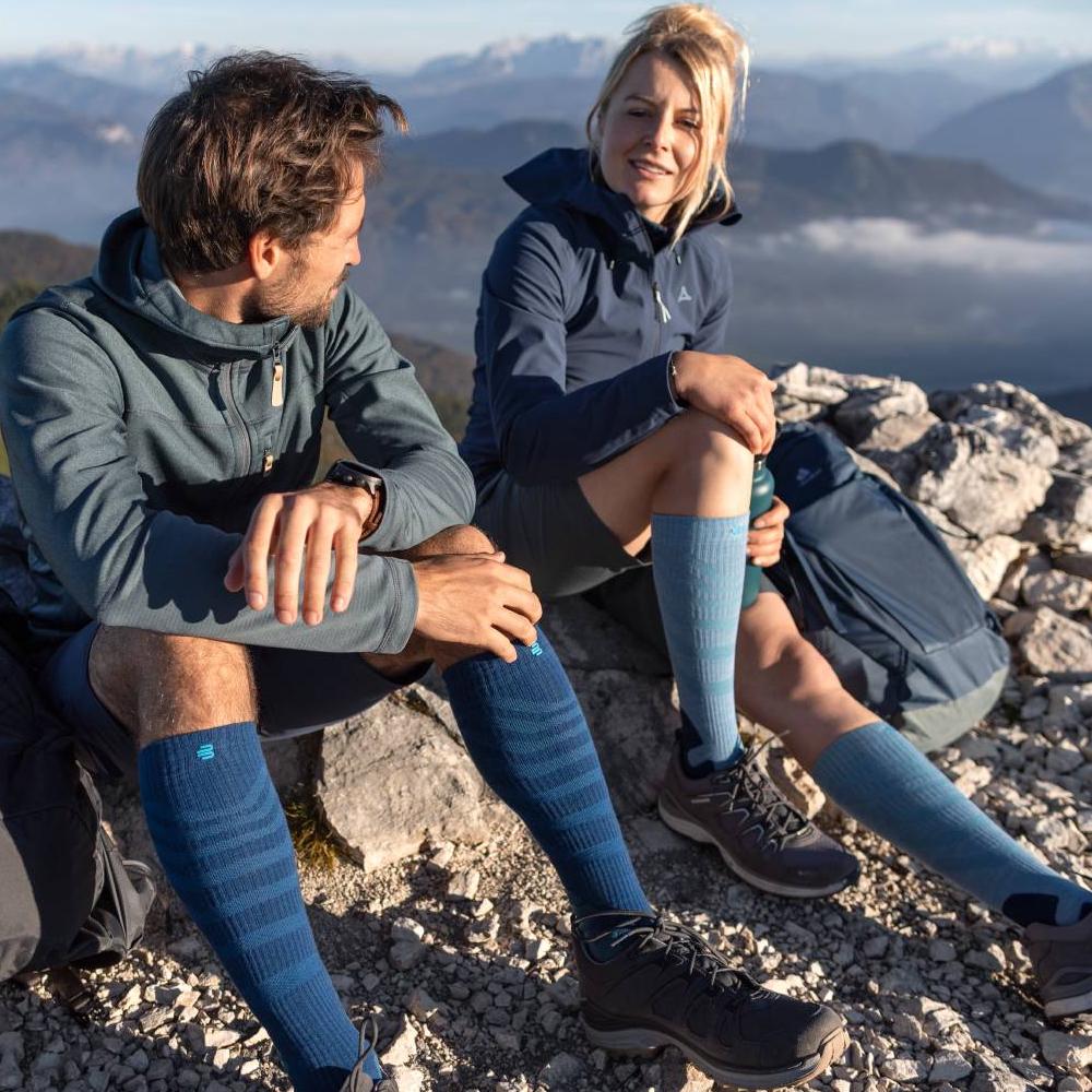 Due escursionisti godono del panorama sulla cima di una montagna, indossando calze a compressione Merino Outdoor in rosso-marrone. Le calze offrono comfort e supporto ottimali per lunghe escursioni in terreni impegnativi.