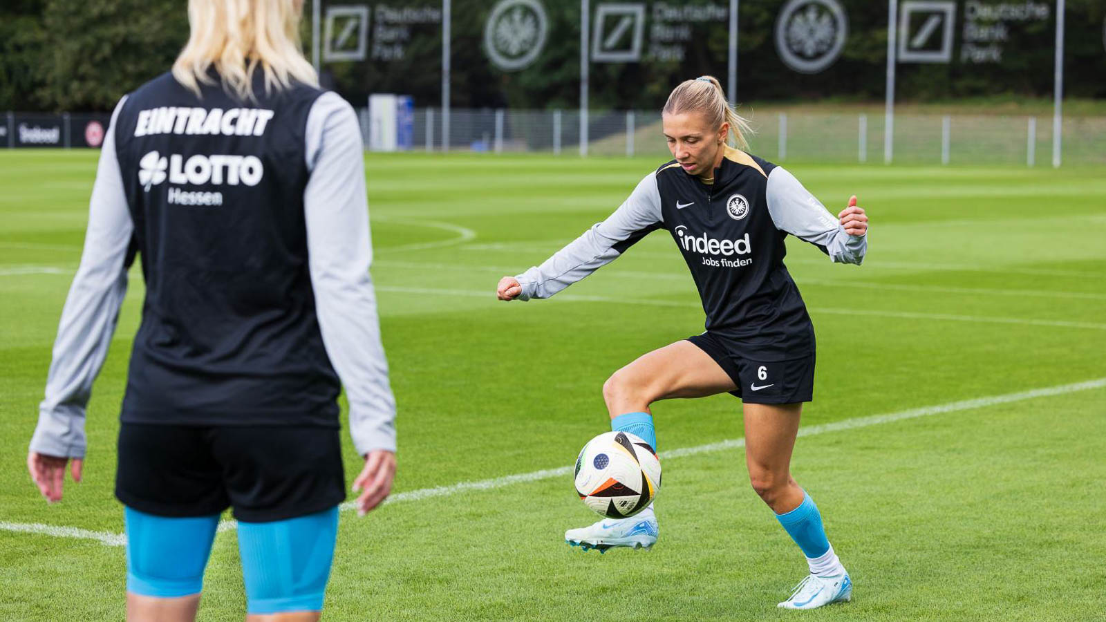 Giocatrice dell'Eintracht Francoforte in azione mentre controlla il pallone. Un'altra compagna osserva l'allenamento sullo sfondo. Focus su tecnica e precisione.