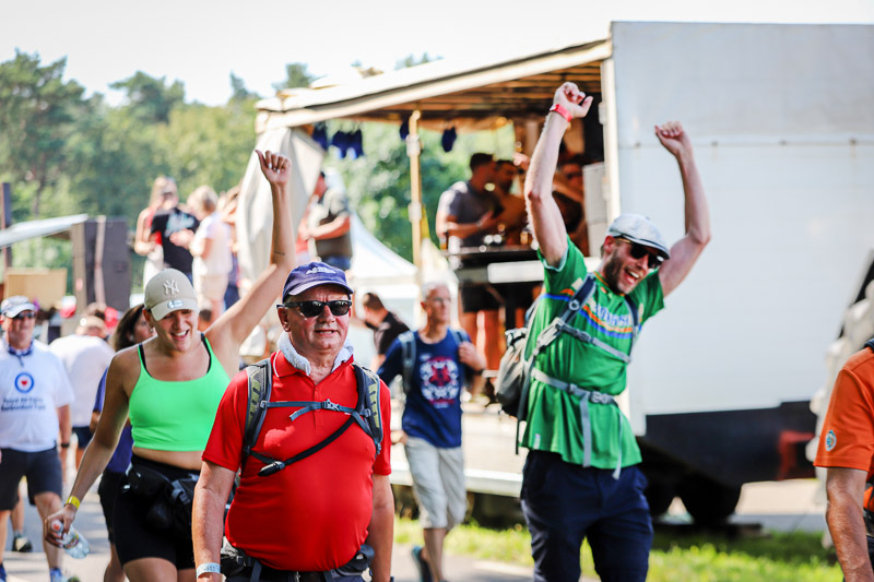 Escursionisti festosi alla 4Daagse Partecipanti felici festeggiano durante la 4Daagse davanti a un palco musicale. Una donna con top verde e un uomo con maglietta rossa passano sorridendo.