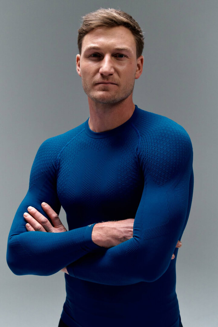 Un uomo con le braccia incrociate indossa una maglia sportiva aderente blu su sfondo grigio. Il tessuto enfatizza la muscolatura e trasmette forza, determinazione e prestazione atletica.