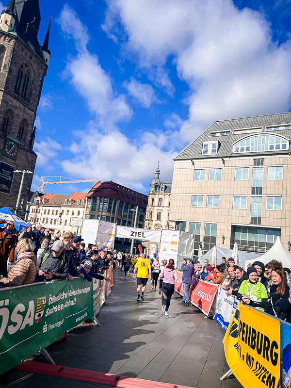 Gli ultimi metri prima del traguardo alla mezza maratona di Halle, con il pubblico che sostiene i corridori Gli ultimi metri prima del traguardo alla mezza maratona di Halle. I corridori attraversano il traguardo sotto un cielo azzurro, incoraggiati dal pubblico.