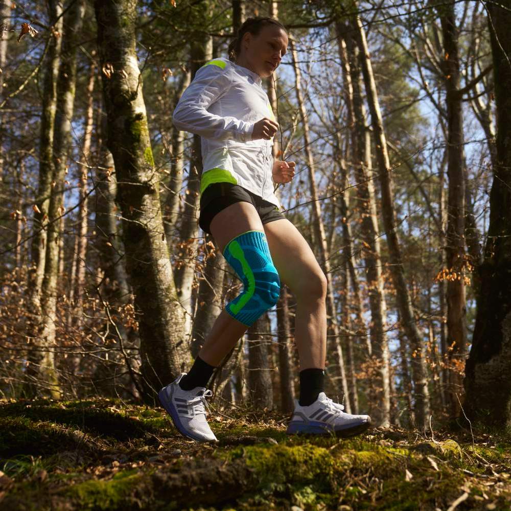 Ginocchiera per il trail running nel bosco Una donna corre su un sentiero nel bosco alla luce del giorno, con una ginocchiera turchese ben visibile sulla gamba sinistra – l’attenzione è rivolta al movimento e all’uso del prodotto all’aperto.