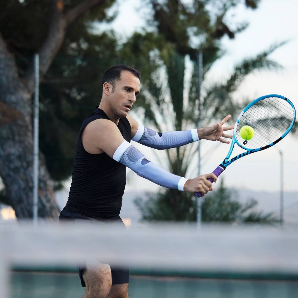 Un tennista con maniche a compressione bianche colpisce un diritto. Sullo sfondo si vedono alberi e un campo da tennis.