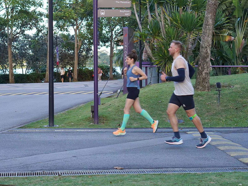 Insieme forti: Allenamento di corsa per la sfida fitness Due persone corrono lungo un parco urbano su una strada asfaltata. Un uomo con abbigliamento chiaro e un tutore al ginocchio corre insieme a una giovane donna con calze a compressione verdi.
