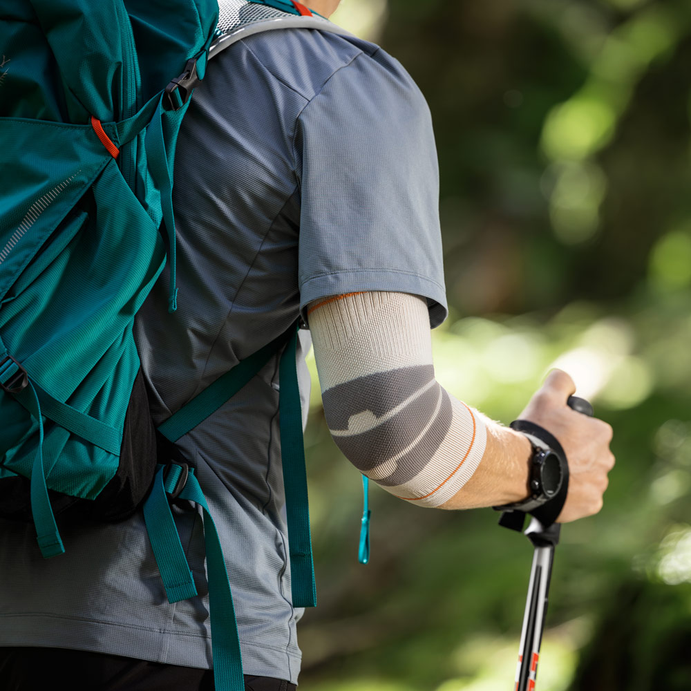 Un escursionista cammina su un sentiero nel bosco utilizzando bastoncini da trekking. Sul braccio destro indossa un Outdoor Compression Elbow Support color sabbia che avvolge l’articolazione del gomito in modo aderente.