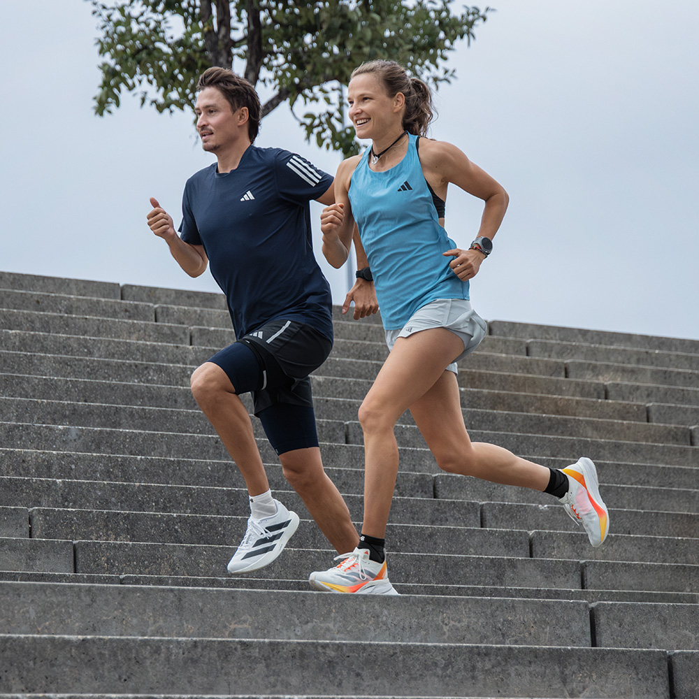In movimento insieme – con agilità giù per le scale Una donna sportiva e un uomo scendono correndo insieme una larga scalinata in pietra. Entrambi indossano abbigliamento sportivo e scarpe adidas.
