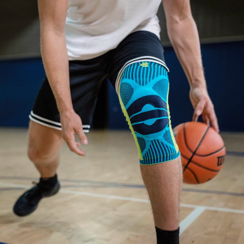 Allenamento di basket con ginocchiera turchese Un giocatore di basket con una ginocchiera turchese sulla gamba destra palleggia in palestra, con enfasi sul prodotto e sulla posizione atletica.