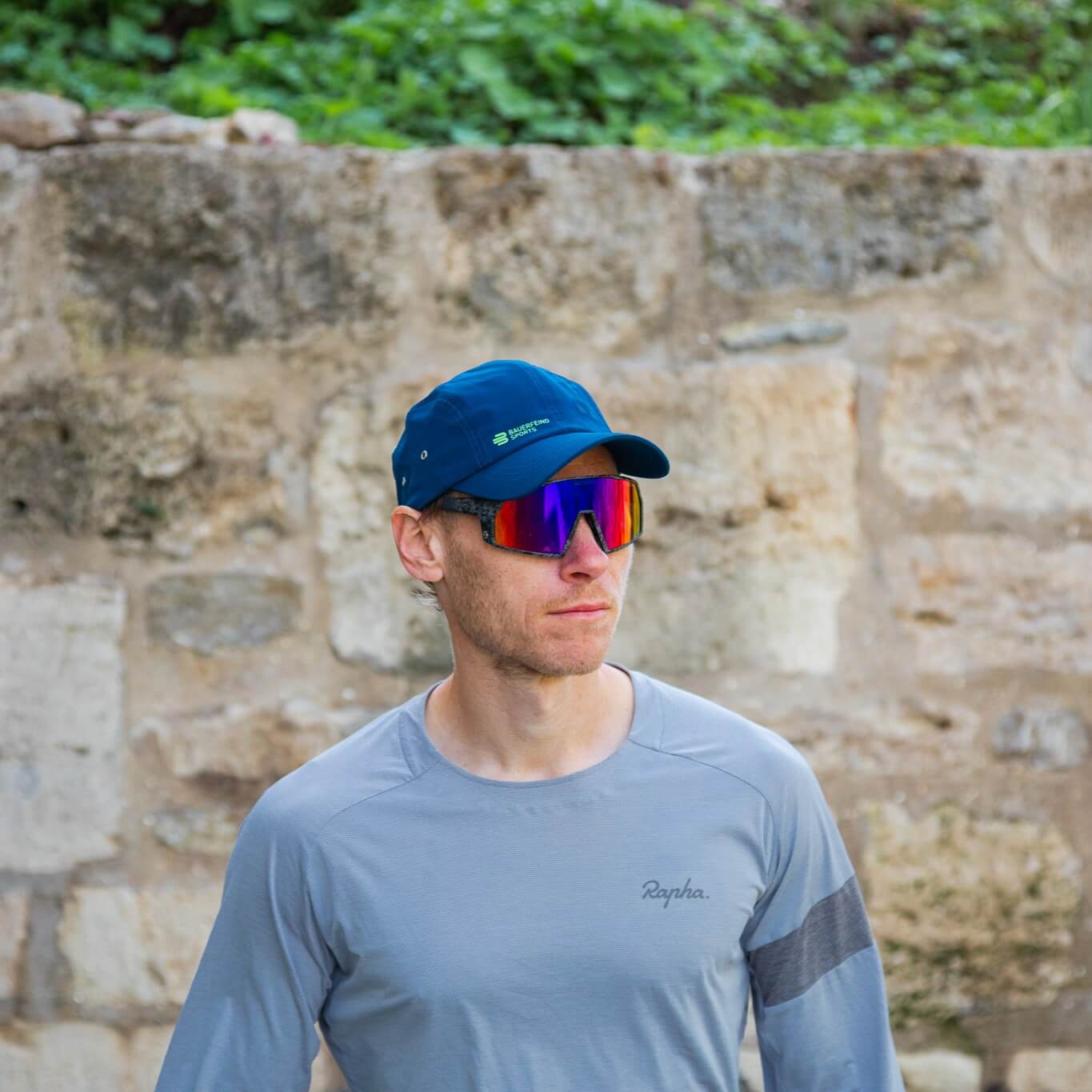 Atleta con cappellino blu Bauerfeind Sports e occhiali sportivi specchiati davanti a un muro in pietra – pronto per vivere lo sport all’aperto.