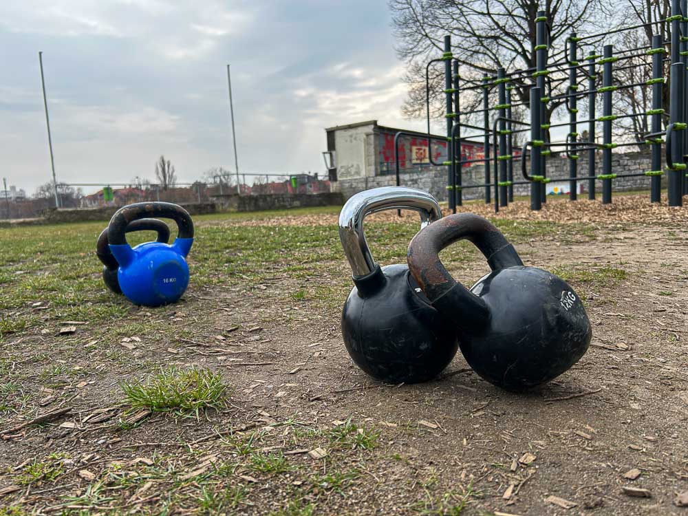 Diverse kettlebell sono appoggiate a terra in un parco outdoor per l’allenamento, con una struttura da calisthenics sullo sfondo. L’immagine mostra un setup funzionale per HYROX senza palestra, ideale per esercizi di forza e carry all’aperto.