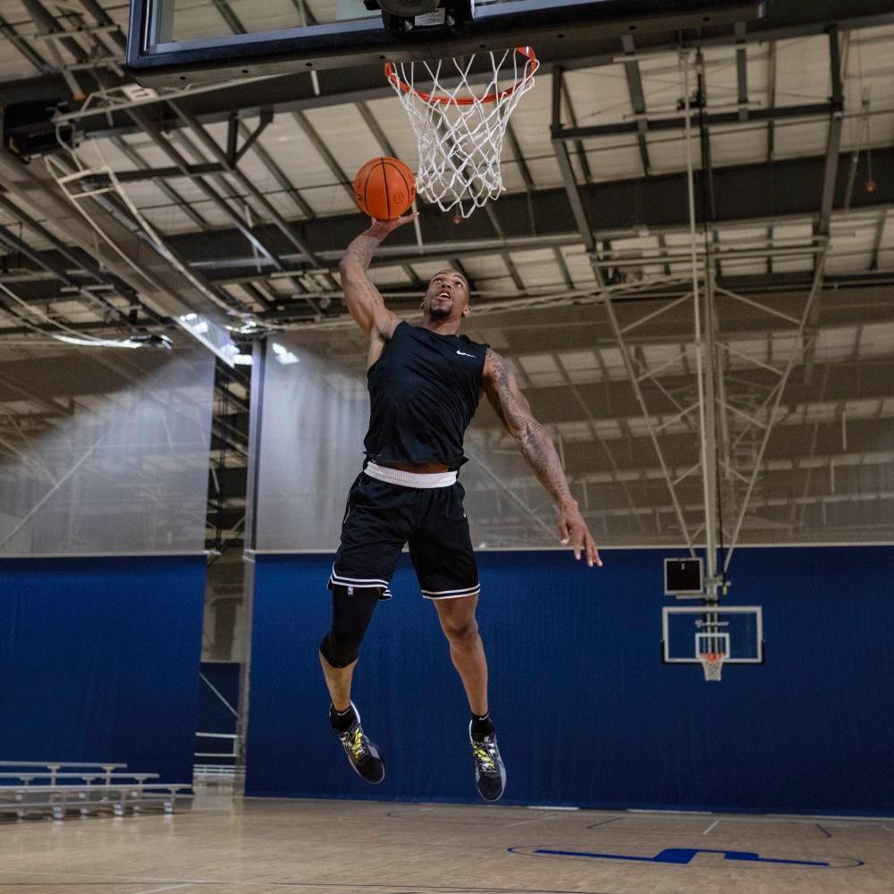 Velocità, abilità, stile – il basket in azione! Un giocatore di basket con una ginocchiera rossa sprinta sul campo all'aperto mentre controlla la palla. Il sole splende e gli alberi sullo sfondo creano un'atmosfera dinamica.
