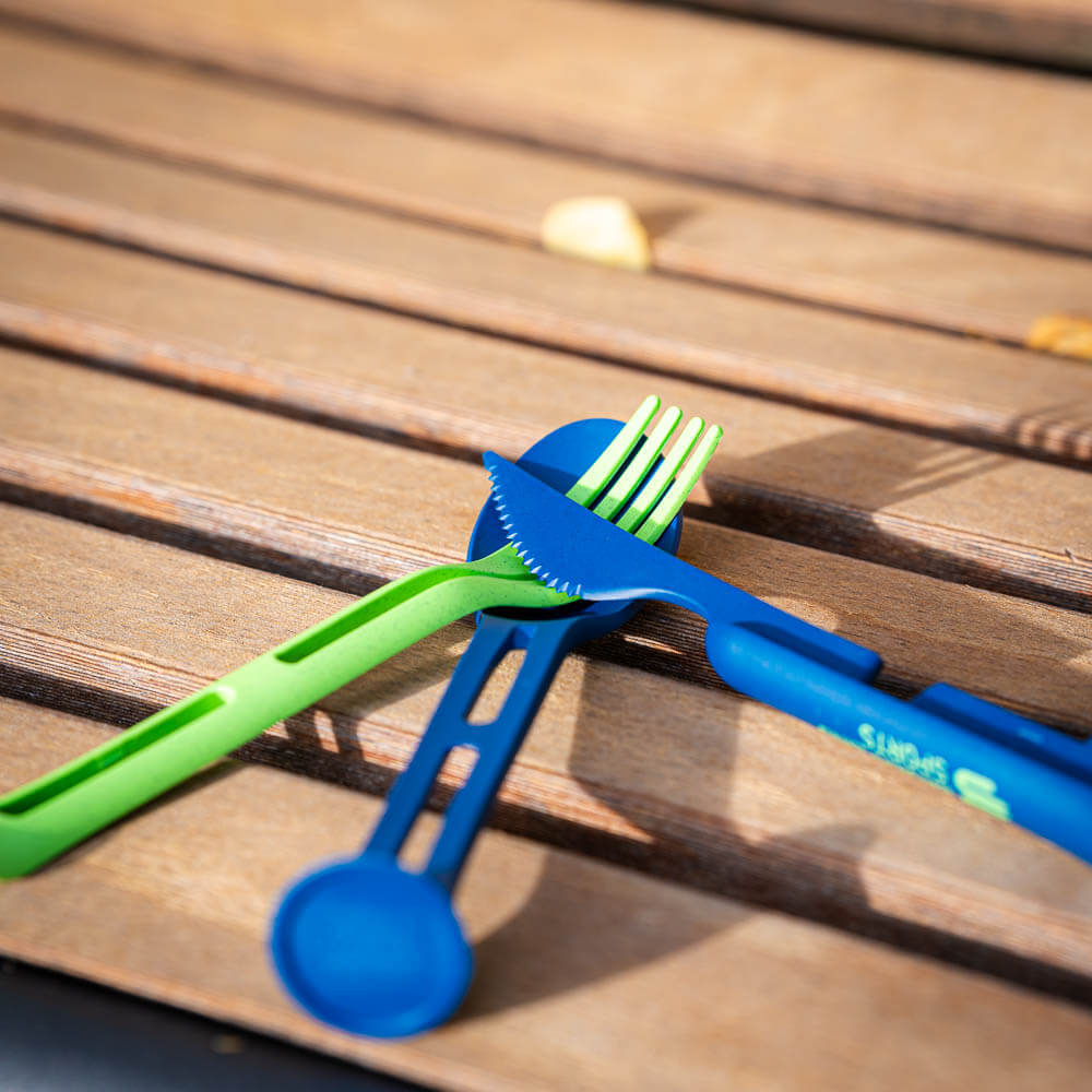 Design intelligente: Set di posate per ogni avventura outdoor Un set di posate verde e blu è incrociato su una panchina di legno. Il set è ideale per pasti all'aperto e confezioni compatte.