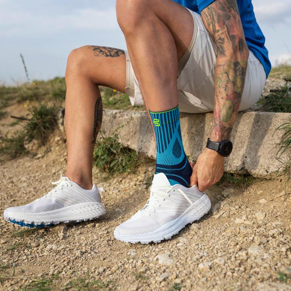 Ein Mann sitzt auf einem Betonsockel in einer natürlichen Umgebung mit Kiesweg und Gras. Er trägt weiße Laufschuhe und blaue Sportsocken mit einem auffälligen Muster. Seine Hände ziehen den Schuh über die Socke, während sein tätowierter Arm sichtbar ist.
