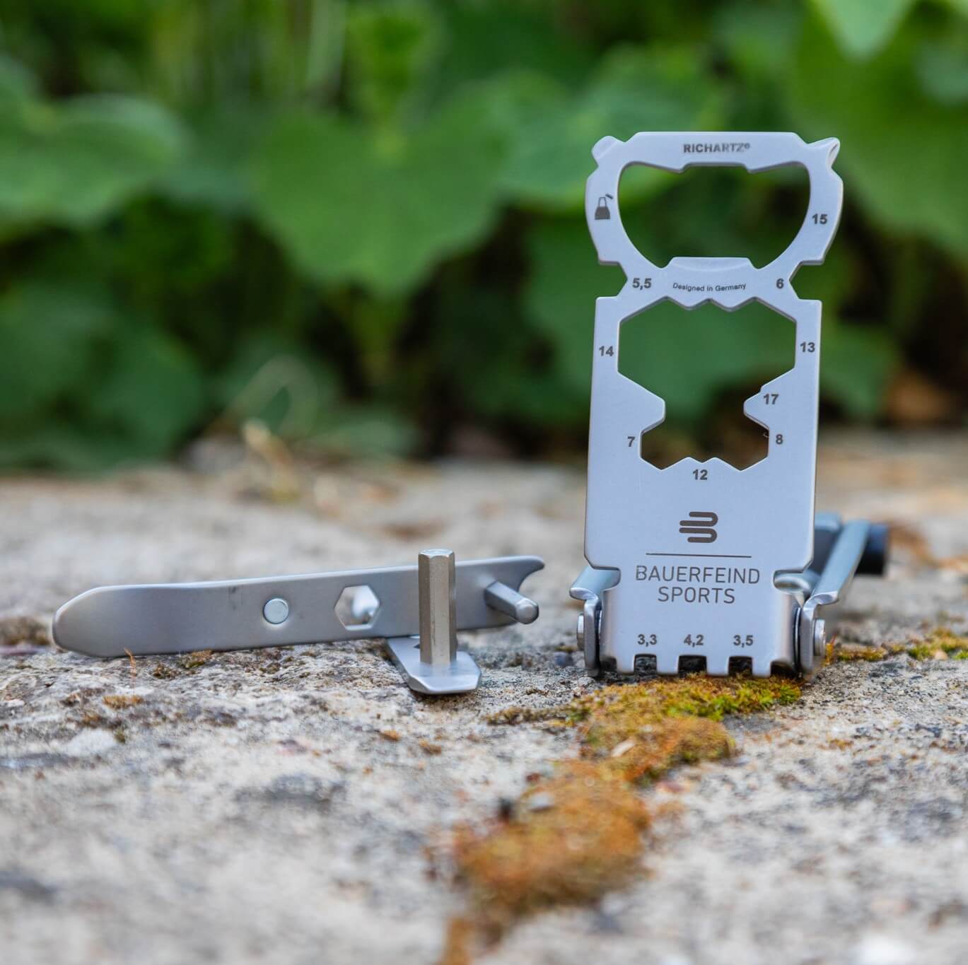 Multitool Bauerfeind Sports con diverse funzioni e una punta separata su una superficie in pietra con muschio, con piante verdi sullo sfondo – lo strumento pratico per riparazioni rapide e uscite in bici più sicure.