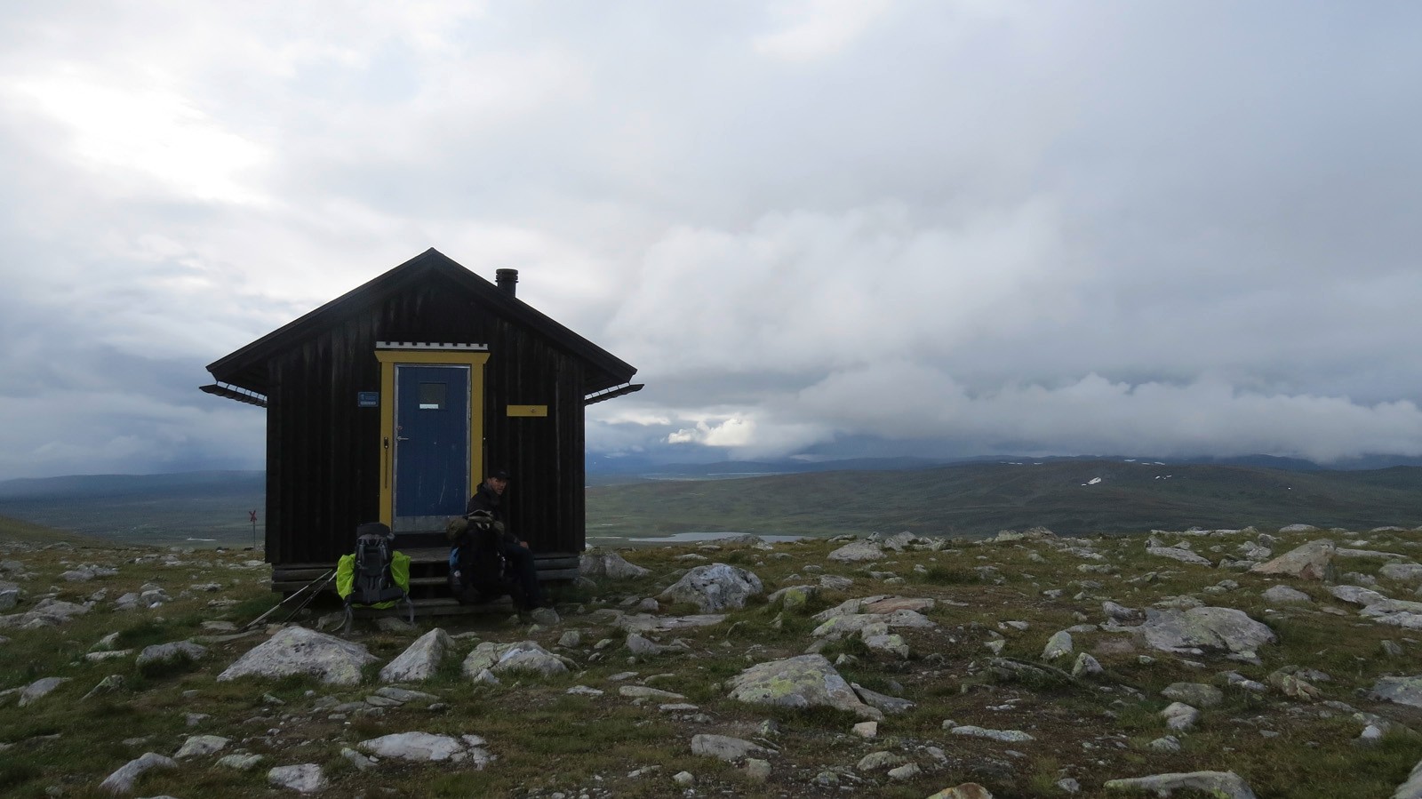 Una capanna solitaria tra le montagne nel mezzo di prati bloccanti con piccole rocce sullo sfondo la fitta copertura nuvolosa e un piccolo lago di montagna