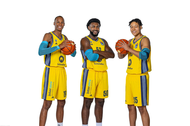 Alba Berlin Team Giocatori – Presentazione Ufficiale Tre giocatori di basket dell'Alba Berlino nelle loro divise gialle posano con palloni da basket davanti a uno sfondo bianco neutro. Ogni giocatore sorride tenendo un pallone con un atteggiamento rilassato e professionale.