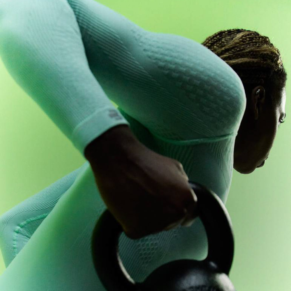 Atleta con top a compressione verde menta e pantaloni a compressione blu scuro che si allena con le braccia Atleta con top a compressione verde menta e pantaloni a compressione blu scuro allunga il braccio verso l'alto mentre tiene in mano un manubrio