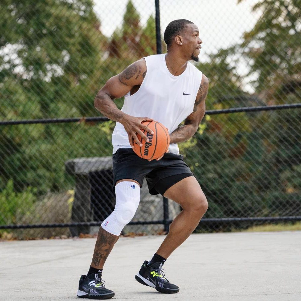 Giocatore con ginocchiera bianca in allenamento al tiro Giocatore di basket con canotta bianca si prepara al tiro. Il ginocchio destro è stabilizzato da una ginocchiera compressiva bianca, sullo sfondo un campo esterno con canestri.