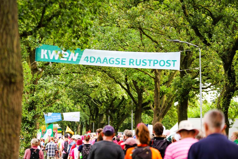 Punto di ristoro alla 4Daagse Numerosi partecipanti passano vicino a un punto di ristoro durante la 4Daagse. Un grande striscione sopra di loro recita "KWBN 4DAAGSE RUSTPOST".