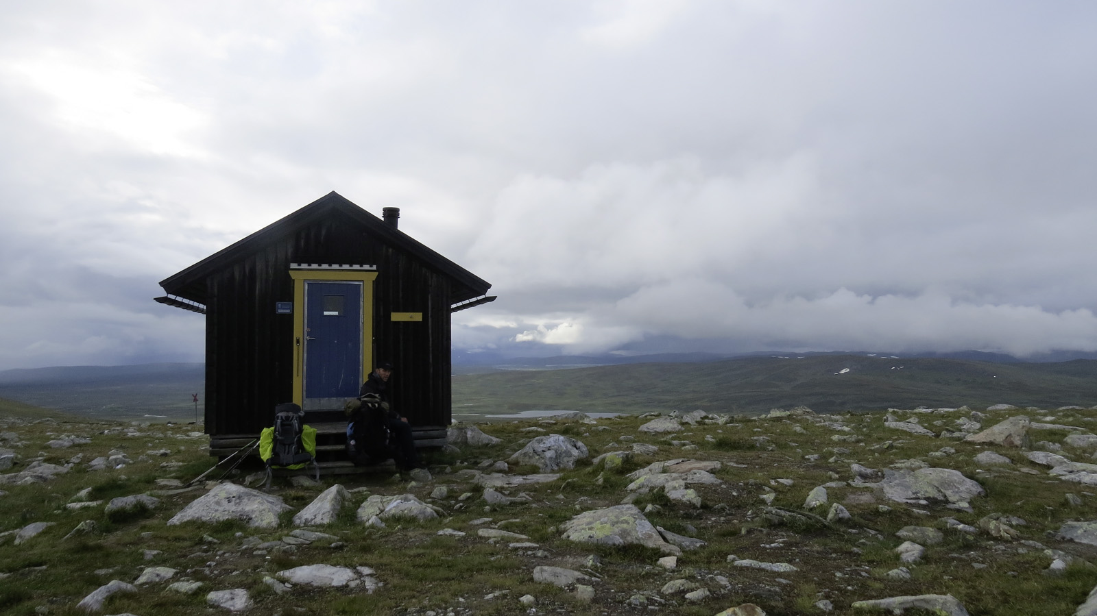 Una capanna solitaria tra le montagne nel mezzo di prati bloccanti con piccole rocce sullo sfondo la fitta copertura nuvolosa e un piccolo lago di montagna
