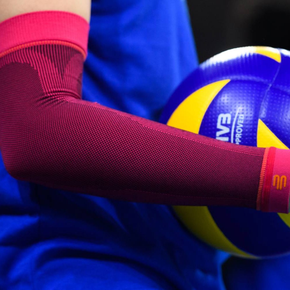 Primo piano di un manicotto da pallavolo rosa sul braccio di un giocatore con un pallone da pallavolo giallo e blu sullo sfondo.