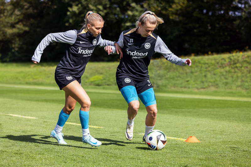 Due giocatrici dell'Eintracht Francoforte lottano per la palla durante una partita di allenamento. Entrambe indossano abbigliamento da allenamento nero con il logo Eintracht e ginocchiere blu di Bauerfeind Sports.