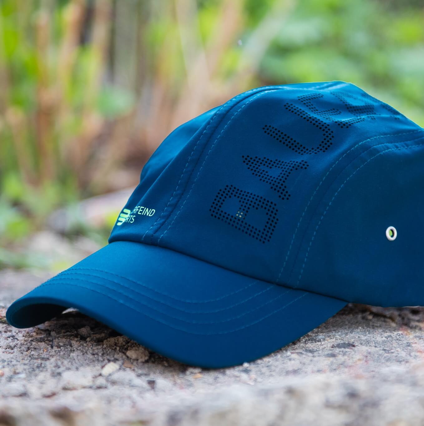 Cappellino blu Bauerfeind Sports visto di lato su una superficie in pietra, con visiera curva, logo verde del brand e scritta BAUERFEIND traforata sul pannello – ideale per vivere lo sport all’aria aperta.