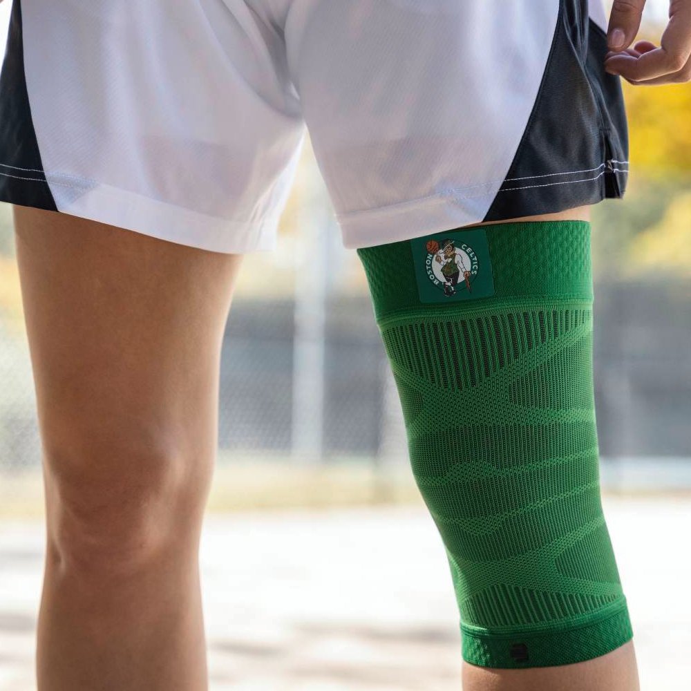 Primo piano di un giocatore con un supporto per il ginocchio verde dei Boston Celtics su un campo da basket. Il supporto presenta il logo della squadra e si adatta ergonomicamente al ginocchio.