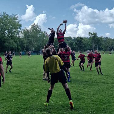 Volare verso la vittoria! Un salto che decide la partita. Una partita di rugby in corso su un campo erboso. Un giocatore con una maglia rosso-nera viene sollevato dai compagni per afferrare la palla, mentre l’arbitro in giallo osserva l’azione.