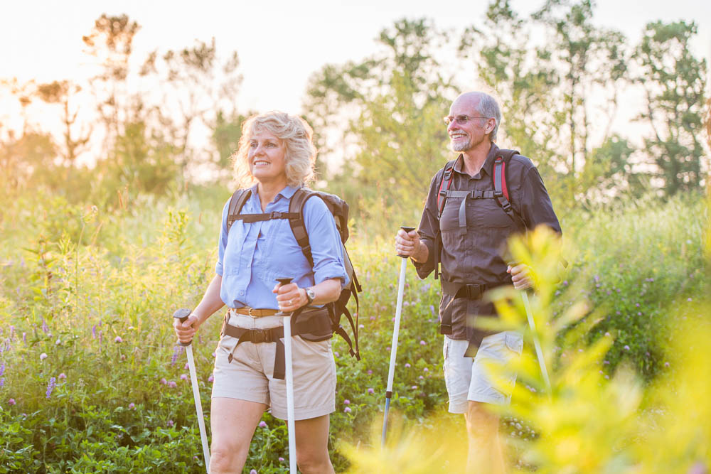Escursione attiva nella luce estiva Una coppia più anziana cammina con bastoncini da trekking attraverso un prato estivo fiorito alla luce del tramonto. Indossano abbigliamento da escursionismo leggero e zaini, e sembrano sereni e attivi.