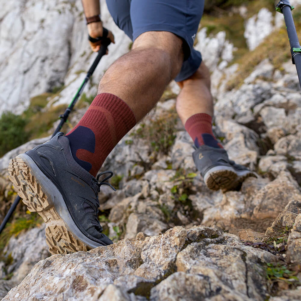 Primo piano di un escursionista che indossa le Merino Outdoor Socks color granato mentre sale su un sentiero roccioso. Le calze offrono supporto e comfort su terreni impegnativi.