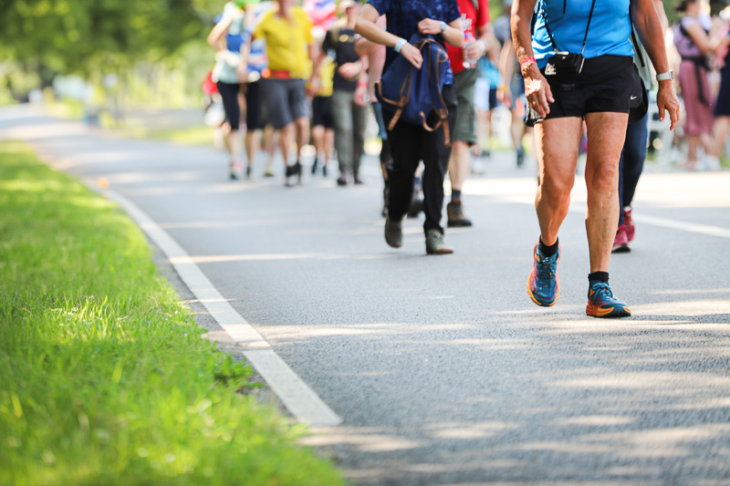 Gruppo di camminatori alla 4Daagse Un gruppo di camminatori percorre una strada chiusa immersa nel verde in una giornata di sole. In primo piano, un uomo con abbigliamento sportivo blu e scarpe da corsa.
