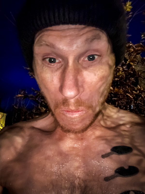 Il grande tuffo: Quando lo sguardo da bagno ghiacciato si fa serio! Un uomo con un berretto nero e il corpo bagnato guarda teso nella fotocamera. Lo sfondo mostra foglie autunnali sotto un cielo serale scuro.