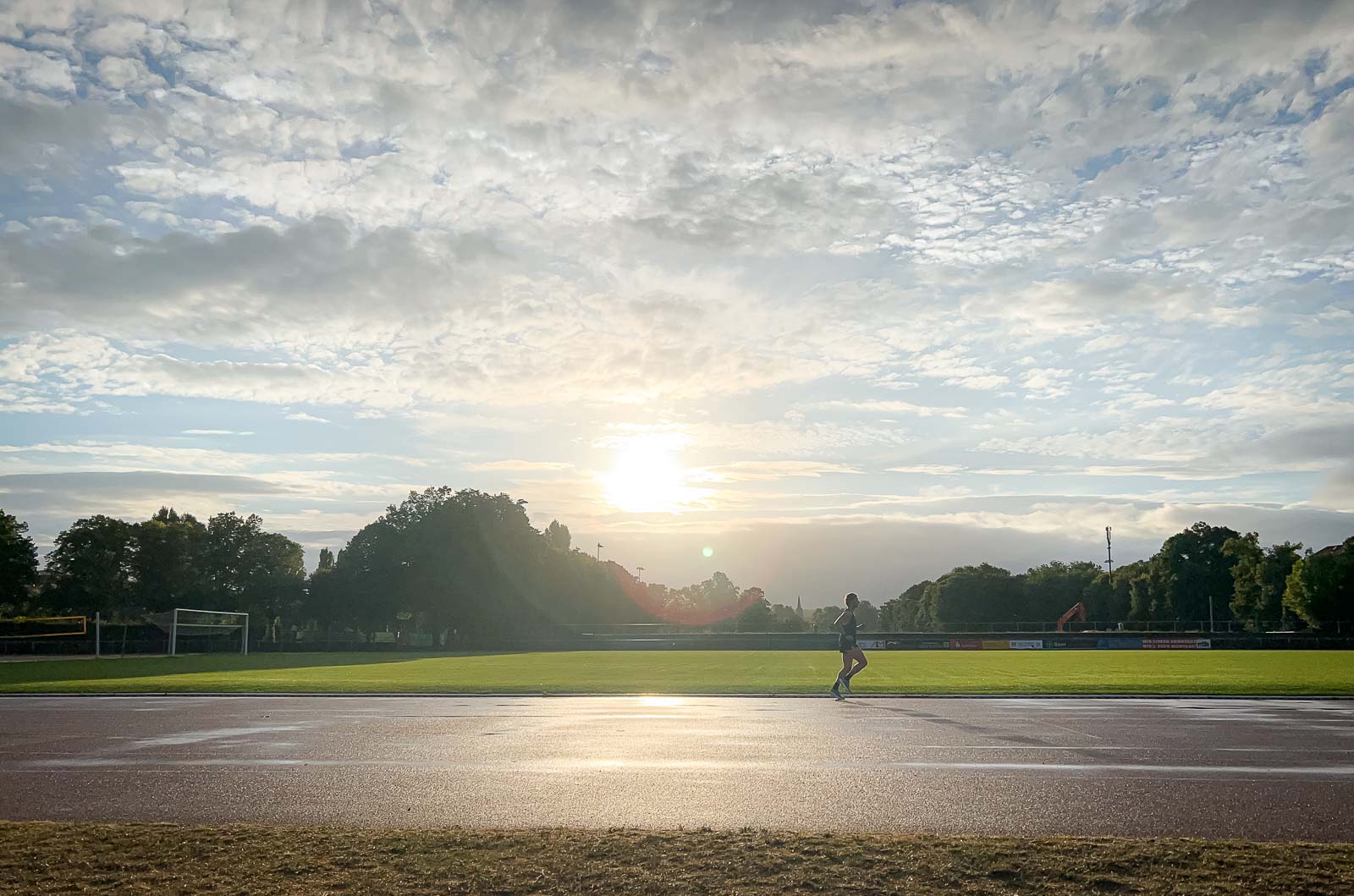 Corridore sulla Pista – Allenamento all'Alba per Resistenza e Velocità. Un corridore su una pista bagnata all'alba. Il cielo è nuvoloso, ma il sole sta facendo capolino mentre il corridore completa il suo giro.