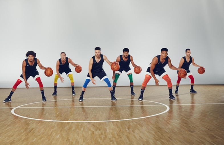 Sei giocatori di basket sono schierati in una palestra, pronti per un'esercitazione. Indossano maglie nere, pantaloncini bianchi e fasce compressive colorate alle ginocchia. Ognuno ha un pallone da basket in mano, pronti per iniziare l’allenamento.