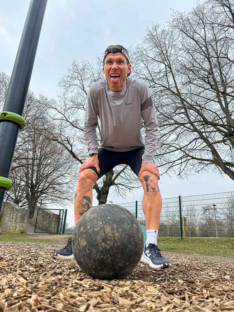 Un atleta si ferma dopo una sessione intensa in un parco outdoor con le mani sulle ginocchia sopra una palla medica. L’immagine mostra la fatica dopo un workout funzionale tipico dell’allenamento HYROX senza palestra.