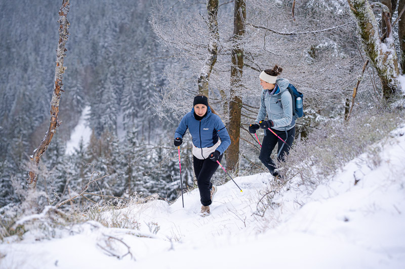 Con i bastoncini nella neve – Escursioni invernali per amanti delle vette! Due donne camminano con bastoncini da trekking attraverso un paesaggio invernale innevato. Indossano abbigliamento tecnico caldo, una giacca blu e una azzurra, guanti e fasce per la testa. Sullo sfondo si vedono alberi innevati e un sentiero ripido.