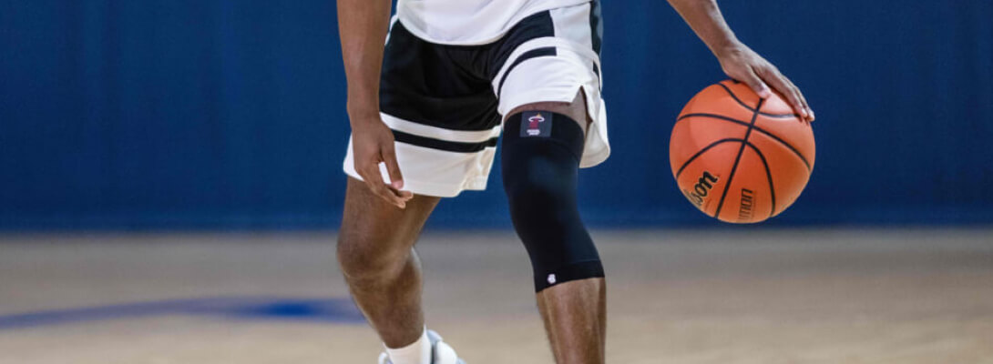 Alles im Griff mit der unserer Compression Knee Support Il giocatore di basket dettagliato con un basket in mano indossa una manica del ginocchio NBA nera
