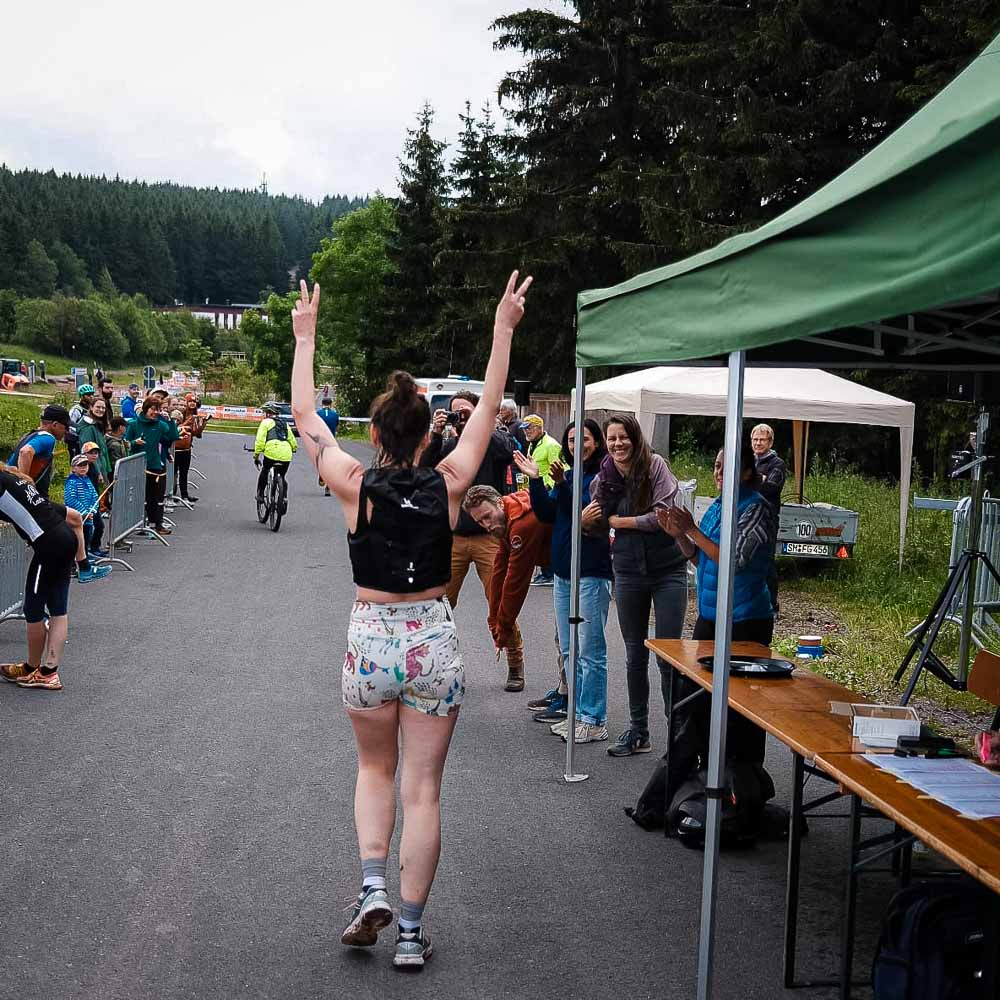 Correre lungo il Rennsteig con il sostegno della squadra e il buon umore Una donna in abbigliamento sportivo fa jogging attraverso il Rennsteig e alza entrambe le braccia per fare il segno della pace mentre gli spettatori la acclamano.