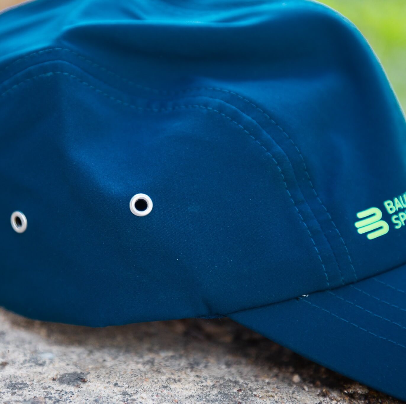 Dettaglio del cappellino blu Bauerfeind Sports con occhielli metallici laterali, cuciture precise e logo verde parzialmente visibile sul pannello frontale – un accessorio sportivo per allenamento, uscite e uso quotidiano.
