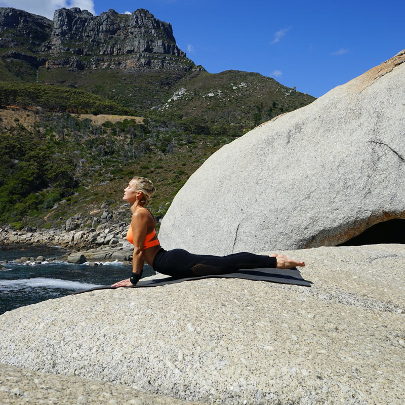 Yoga all'aperto - Posa di Königskobra Donna con bendaggio da polso sulle rocce nella posa del re Cobra sostiene i suoi palmi sulle gambe dritte sul pavimento