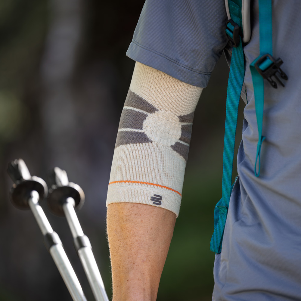 Un escursionista cammina su un sentiero nel bosco utilizzando bastoncini da trekking e indossa un Outdoor Compression Elbow Support color sandstone sul braccio destro.