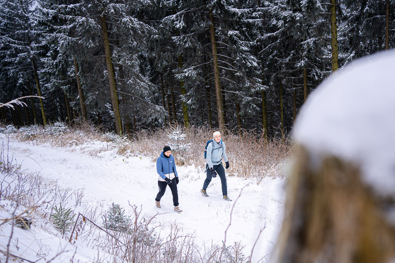 Escursione invernale – Dove ogni passo è un'avventura scricchiolante. Due donne camminano attraverso un paesaggio boschivo innevato. Indossano abbigliamento invernale e utilizzano bastoncini da trekking mentre percorrono un sentiero. Sullo sfondo si vedono alberi ricoperti di neve e una foresta fitta.
