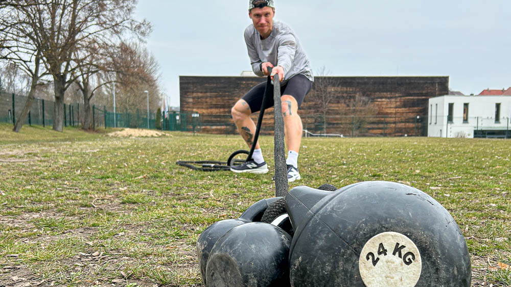 Atleta trascina due kettlebell con una cinghia sull’erba durante un allenamento HYROX all’aperto: forza funzionale senza palestra.