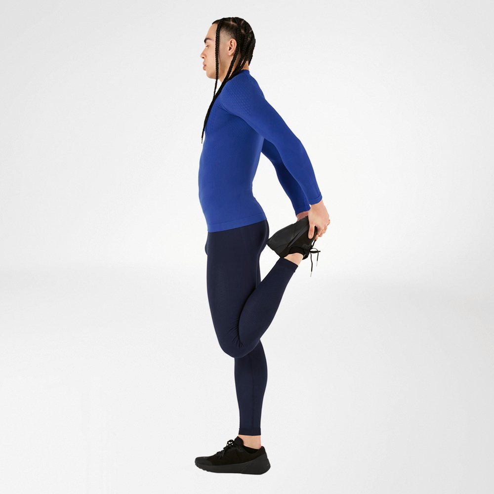 Un atleta indossa un abito a compressione blu, composto da una maglia a maniche lunghe e collant, esegue un esercizio di stretching, tenendo una gamba indietro, su uno sfondo bianco. L'abito offre un alto comfort e un supporto muscolare ottimale.
