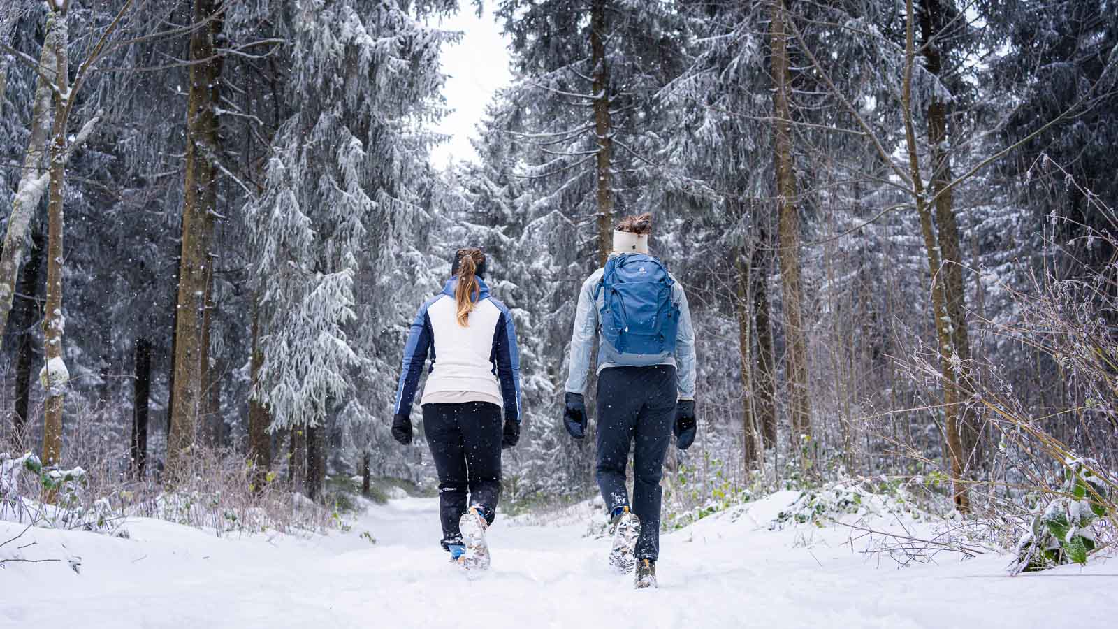 Magia invernale: Segui il sentiero verso l'avventura bianca Due donne camminano attraverso una foresta coperta di neve, viste da dietro. Indossano giacche invernali, scarponi e guanti. Una delle donne ha uno zaino blu. Il sentiero innevato si snoda tra abeti carichi di neve.