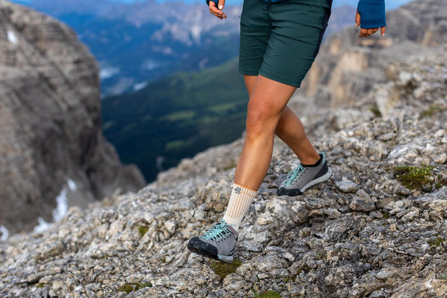 Un escursionista si muove su un terreno roccioso con scarpe da outdoor e una cavigliera arancione. Sullo sfondo si estende un vasto paesaggio montano.