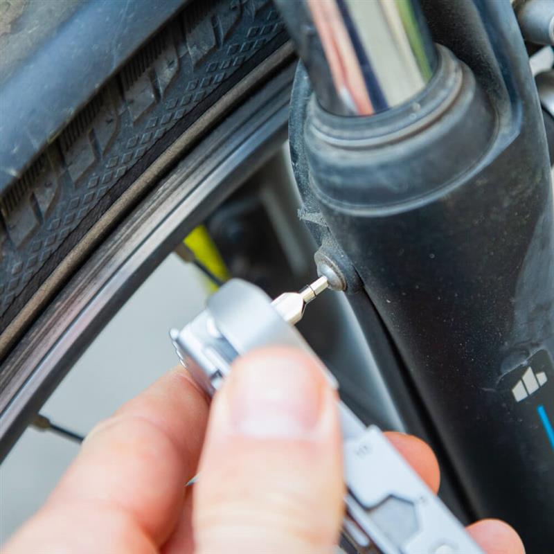 Primo piano di una ruota e della forcella della bici, con una mano che utilizza un multitool grigio Bauerfeind Sports per regolare con precisione una piccola vite – per una bici affidabile e più sicurezza in ogni uscita.